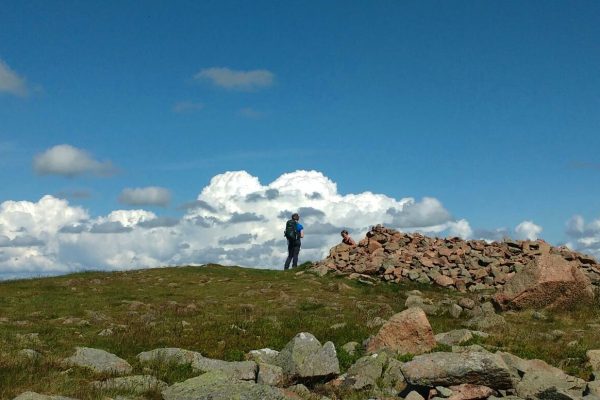 A walker on a hill top with clouds in the distance and a cairn to the right