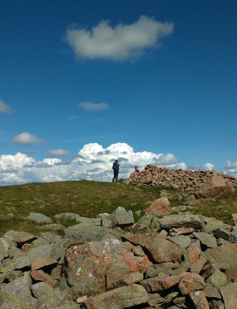 A walker on a hill top with clouds in the distance and a cairn to the right