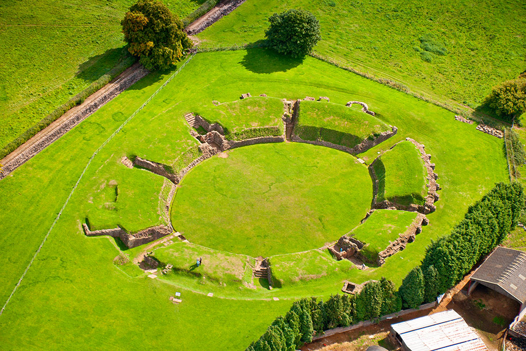 Grassy field with remains of an amphitheatre. A level central circle can be seen, surrounded by eight raised segments forming a ring around it.