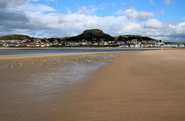 View across a beach to a town below a prominent hill surrounded by cliffs